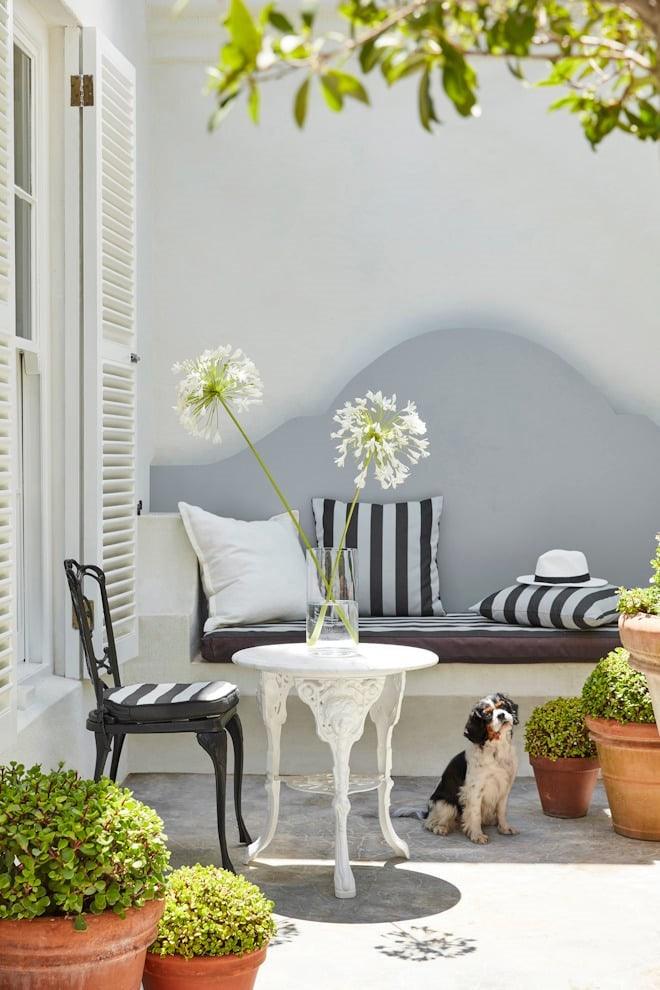 Helle Vintage Gartenlounge mit weißen Möbeln, schwarz-weißen Kissen, Grünpflanzen und kleinem Hund